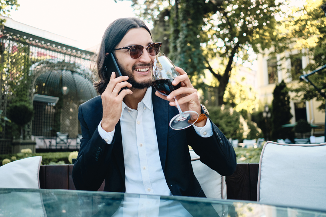 Young handsome smiling brunette bearded man happily talking on cellphone and drinking wine resting in restaurant outdoor