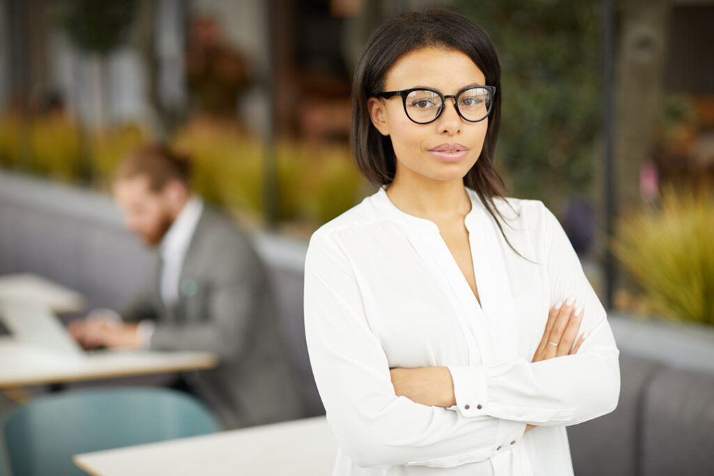 Portrait of confident young businesswoman in eyeglasses and in white blouse standing with arms crossed and looking at camera with man in the background