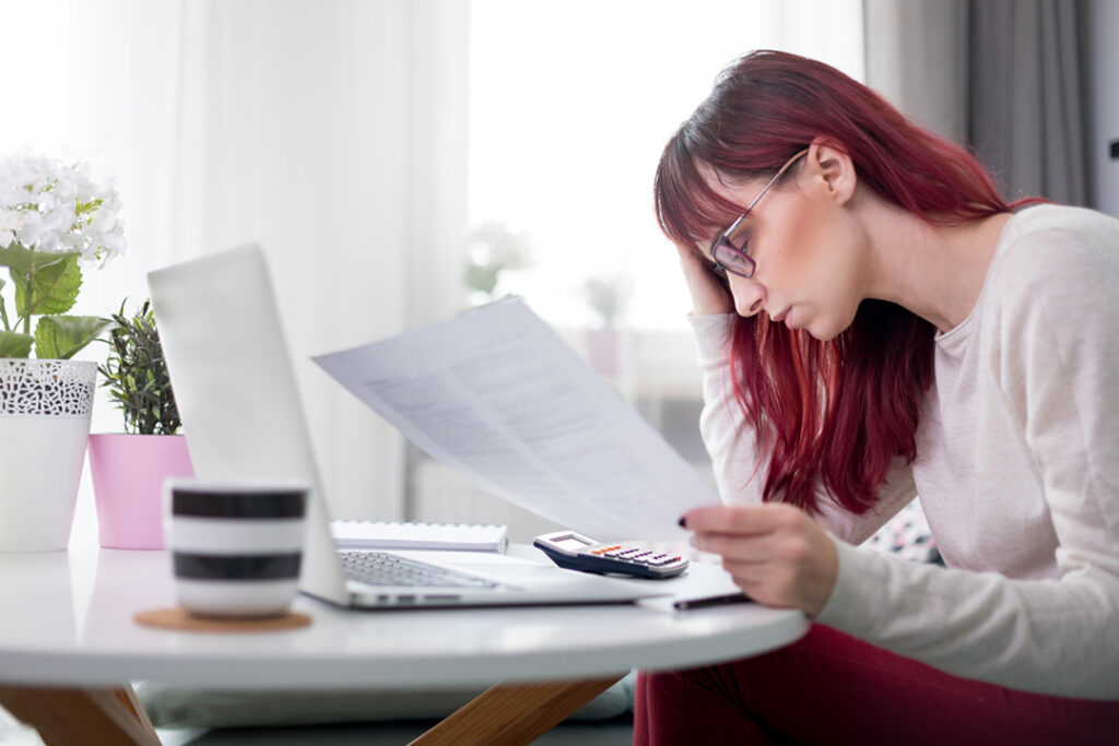 Worried woman at home checking financial documents using calculator and laptop