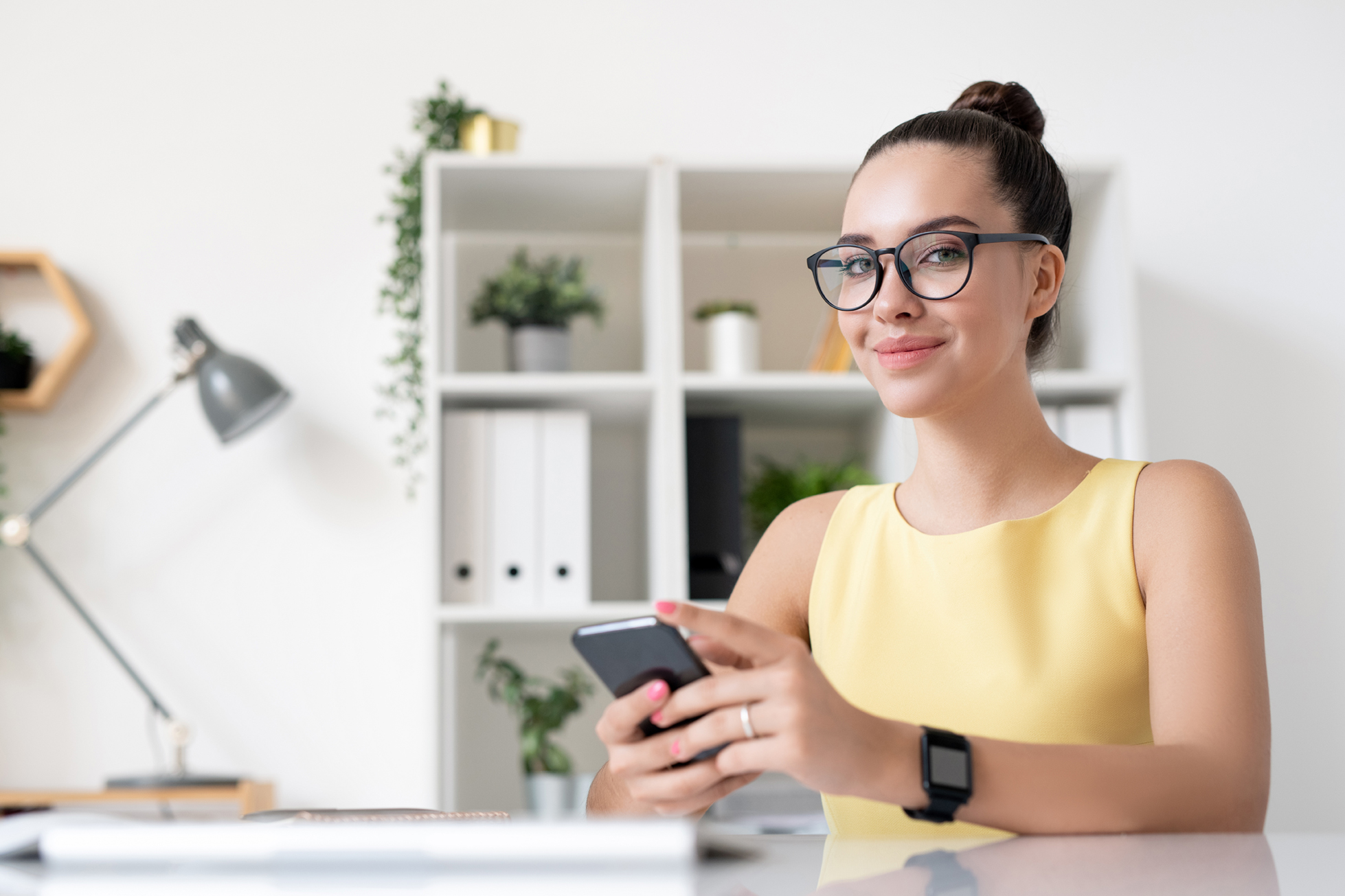 Portrait of content young marketing specialist in yellow dress sitting with smartphone in modern office